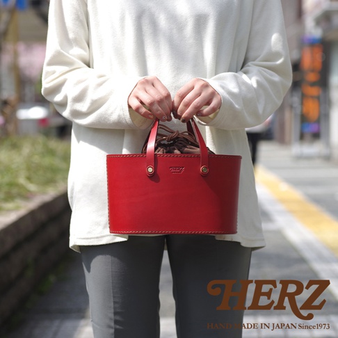 職人手作紅色皮革束口手提包，帶有深棕色束繩和同色系皮革提把，包身結構方正，縫線清晰可見，低調的品牌壓印在包面上。