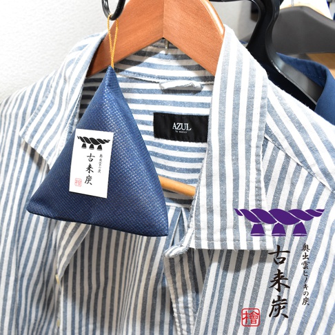 一件懸掛在木製衣架上的藍色三角形麻布包，包裝正面附有寫著古來炭字樣的白色標籤，圖案為深紫色注連繩，背景是一件藍白條紋的AZUL襯衫，整體呈現出簡約的居家除濕與除臭氛圍。