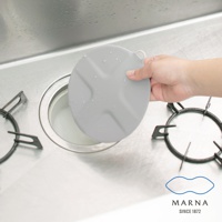 灰色圓形矽膠水槽蓋，表面有十字狀壓紋，邊緣帶有一個小掛環，一隻手正將其放置在廚房水槽排水口上。