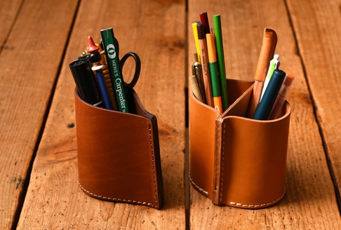 兩個手作皮革分隔筆筒，皆為淺棕色，左邊筆筒內有筆、剪刀等文具，右邊筆筒內有彩色鉛筆、簽字筆等文具。