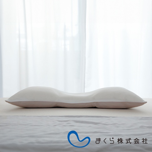 無敵低枕，白色網狀表層，內襯淺褐色布料，呈現S型曲線設計，置於白色床單上，後方為白色紗簾。