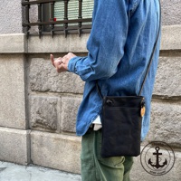 黑色帆布皮革散步包，配有可調節肩帶和外部口袋。包包上有一個圓形吊牌，上面印有船錨圖案和「clueto KOBE since 2001」字樣。