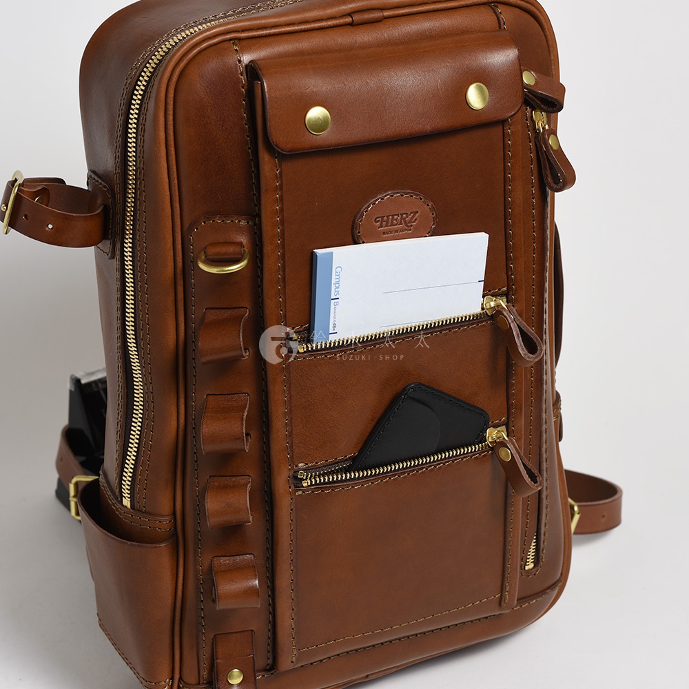棕色皮革製成的職人手作兩用後背包，設有拉鍊主隔層、前置翻蓋口袋、兩個拉鍊前袋，其中一個袋內可見筆記本及手機，並有側袋及皮革提把。