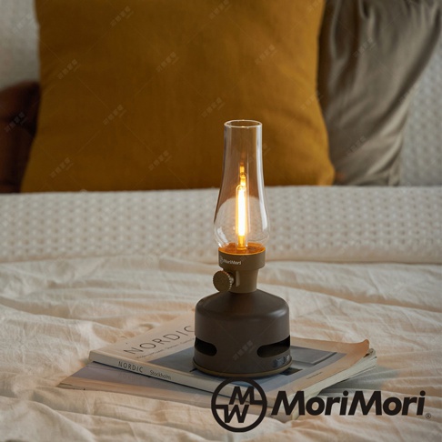 深棕色煤油燈造型藍牙音響，燈罩為透明玻璃，內置暖黃色LED燈條，底部有圓形喇叭孔和旋鈕，置於雜誌上。