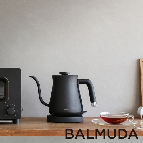 黑色啞光質感的 BALMUDA The Pot 手沖壺，壺身呈圓潤的水滴狀，搭配細長的彎曲壺嘴和人體工學手把。壺身側面印有 BALMUDA 標誌，置於同色系黑色底座上。