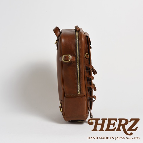 職人手作皮革兩用多功能後背包 R-66，展示的是一款棕褐色皮革後背包的側邊視角。包身由光滑皮革製成，配有金色拉鍊、扣環和D型環。包側有數個皮革繫帶。