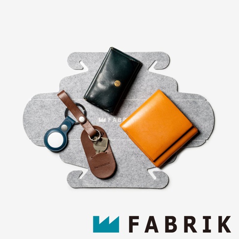 FABRIK 品牌標誌旁，展示著數個皮革小物，包括一個深綠色短夾、一個橘黃色錢包、一個皮革鑰匙包，以及一個附有藍色保護套的物品追蹤器。