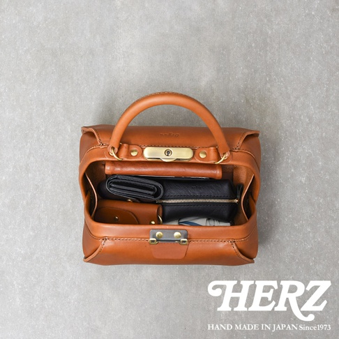 職人手作皮革Organ兩用口金手提包，採用天然皮革製成，呈現溫潤的焦糖棕色。包款設計簡潔，有手提把和金屬鎖扣，內部可見皮夾、零錢包等物品，展現其收納功能。