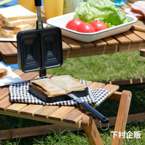 直火式壓邊吐司烤盤，黑色，帶有兩格烤盤，烤盤內有兩塊已烤好的吐司。烤盤放置在木製折疊桌上，桌上還有生菜、番茄和其他食材。