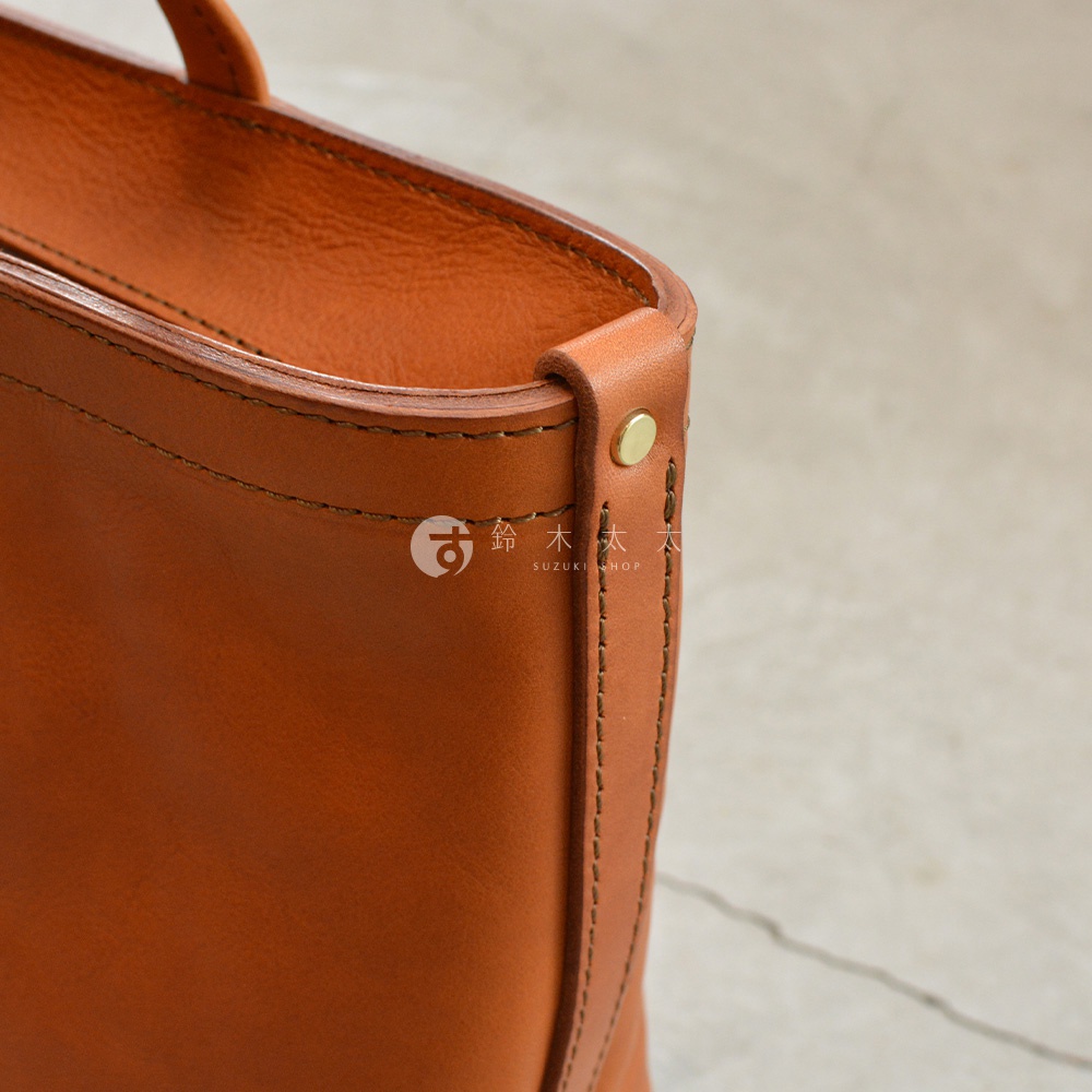 職人手作皮革包的細節，包含縫線、金屬鉚釘與皮革邊緣，以及上方浮水印「鈴木太太 SUZUKI SHOP」。