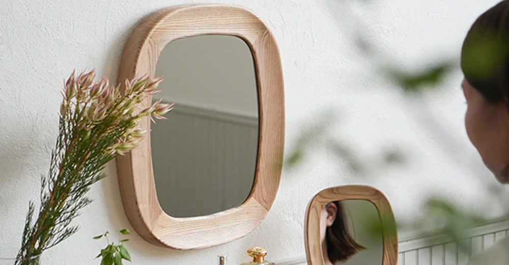 懸掛在白色牆面上的 MOI 質感木框萬用壁掛鏡，擁有圓潤邊角與淺木色質感的木製鏡框，鏡面為橢圓形，映照出旁側的乾燥花束與遠處的小鏡子。