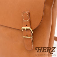 日本HERZ職人手作皮革雙扣旅行郵差包，展示包袋的銅扣和皮革紋理，下方有HERZ品牌標誌及MADE IN JAPAN Since1973字樣