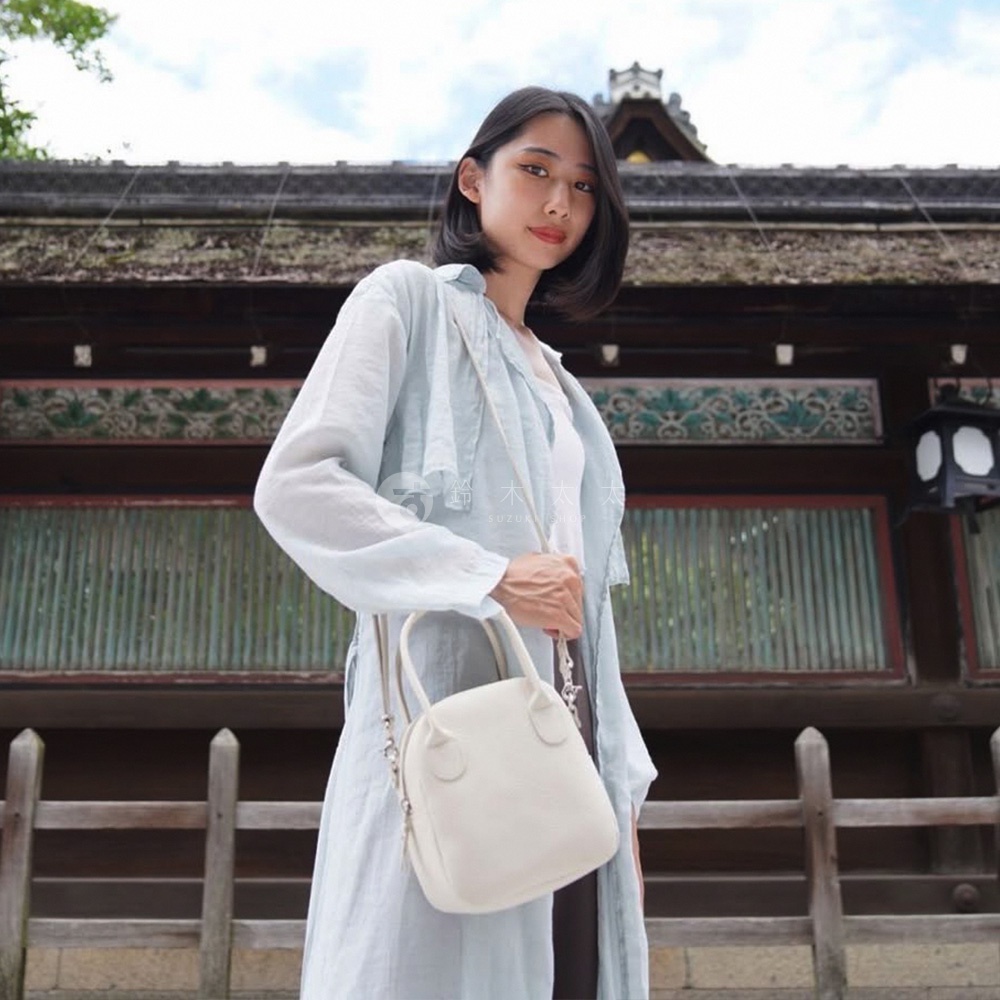 一位女性身穿淺藍色長袍，斜挎一個米白色皮革貝殼包，包包有兩個短提把和一條可拆卸長背帶，背景為日式建築。