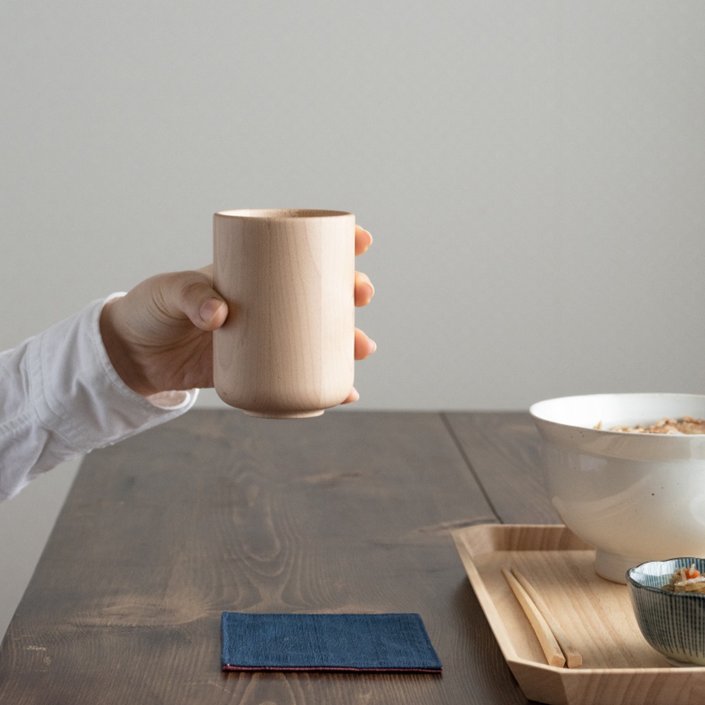 手持一個淺色楓木水杯，杯身圓潤，無把手設計。桌上擺放著藍色杯墊、木質餐盤、一碗粥和一小碟配菜。