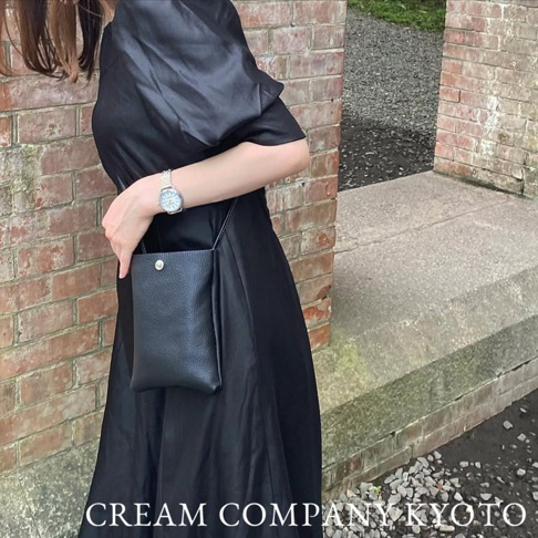 黑色荔枝紋皮革手機包，附有金色按扣，搭配黑色連衣裙，由身著銀色手錶的女性展示。