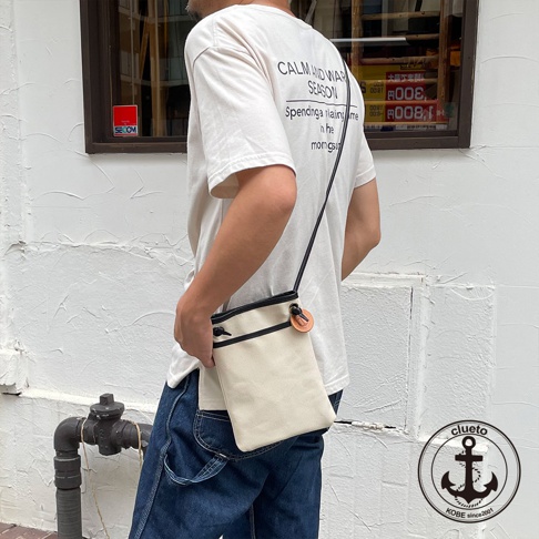 米白色帆布與黑色皮革拼接的斜背包，配有木質鈕扣和束繩開口，由男性身穿白色T恤和牛仔褲展示。