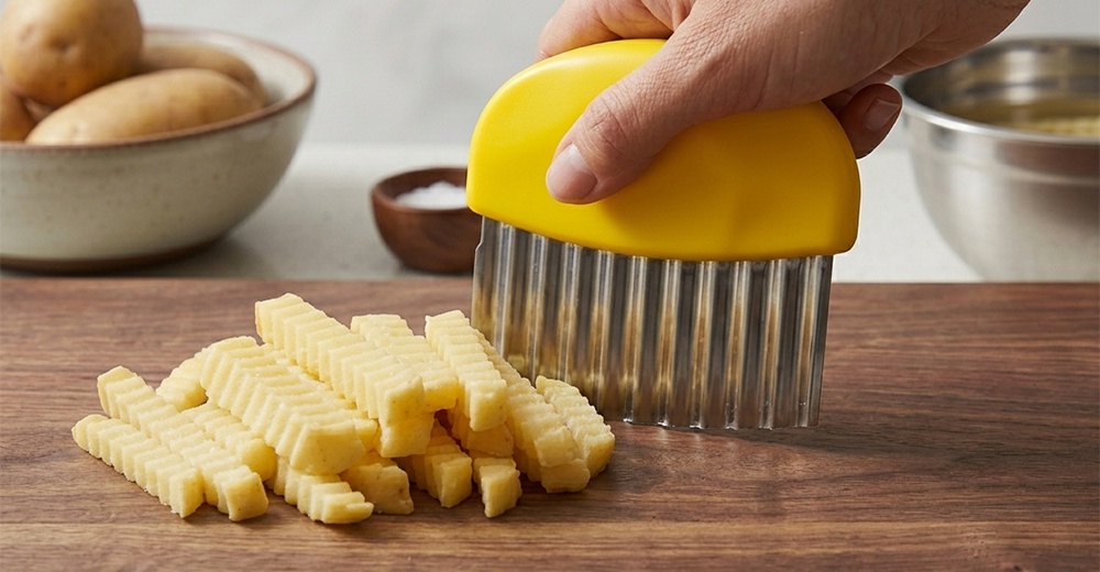 一隻手握住一把黃色手把的波浪造型切刀，正在木頭砧板上切割馬鈴薯，旁邊散落著已經切好的波浪狀薯條，背景設有盛裝馬鈴薯的碗與一個金屬盆。