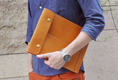 職人手作皮革信封式雙釦手拿袋，採用光滑的棕褐色皮革製成，配有兩個金色釦子和白色縫線細節，由一位穿著藍色襯衫和橘色褲子的人士持握。