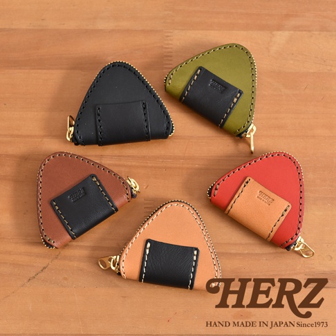 五個不同配色的日本HERZ職人手作皮革三角飯糰造型零錢包，放置在木質桌面上，每個零錢包皆採用對比色拼接設計並配有拉鍊，右下角印有HERZ品牌標誌及產地說明。
