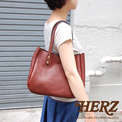 一位女性背著一個棕褐色皮革托特包，包包為開放式開口，有兩條皮革提把，上面有金屬鉚釘裝飾，包身側面有縫線裝飾。