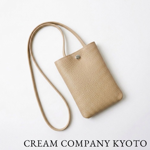這是一款 CREAM COMPANY KYOTO 的日本職人手作隨身手機包，選用質感柔軟且輕量耐用的淺米色牛皮革製成，表面具備細緻紋理，簡約的單一開口設計搭配金屬扣環與細長背帶，適合收納手機及隨身小物。