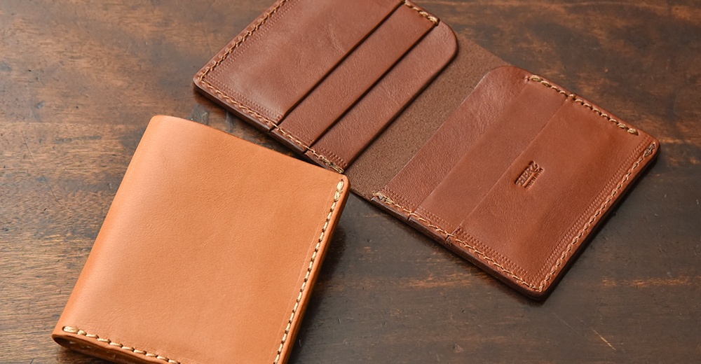 一張木質桌面上擺放著兩個風格簡約的皮革製品，一個為深棕色、已攤開的皮夾，內有三個卡槽；另一個為淺棕色、閉合狀態的皮夾，放置在左側。兩個皮夾都帶有手工縫線，邊緣呈現精緻的職人風格，其中攤開的皮夾內有「HERZ」的壓印品牌標誌。