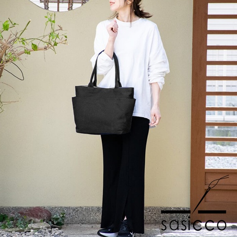 一款黑色三河木綿刺子織手提托特包，包身有兩個外側口袋，搭配白色長袖上衣和黑色長褲。