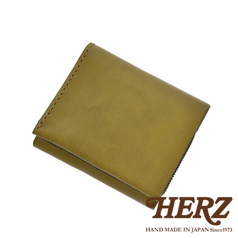 這是一個日本HERZ品牌的手工職人短夾，採用橄欖綠色系的Stare牛皮製作，皮面呈現自然細膩的皮革紋理。錢包外觀簡約俐落，邊緣以細緻的縫線工藝固定，整體呈現出沈穩且充滿質感的職人手作風格，右下角印有深褐色的HERZ品牌標誌與創立年份。