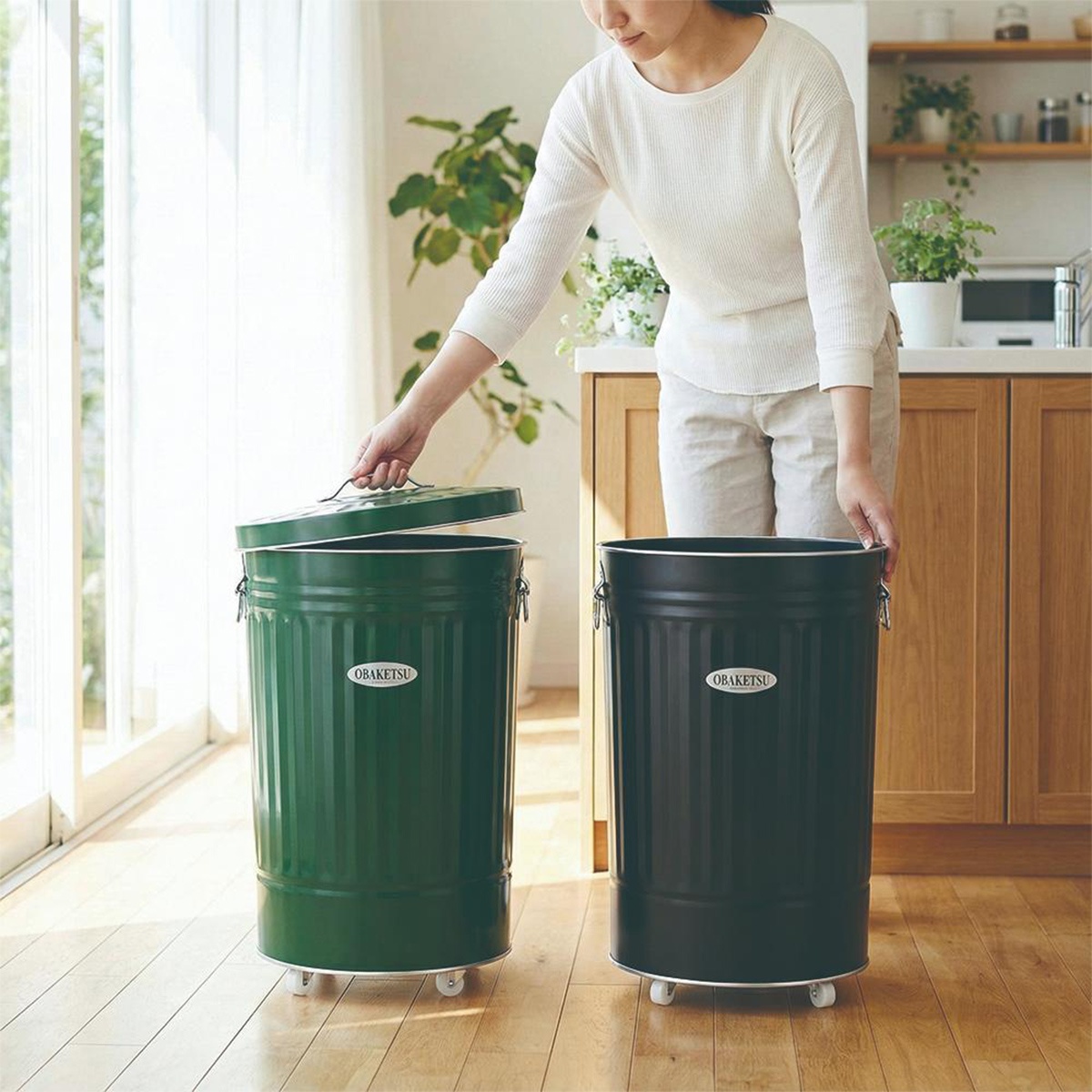 一位女性正在將附輪的OBAKETSU綠色垃圾桶蓋子掀開，旁邊還有一個黑色的OBAKETSU垃圾桶