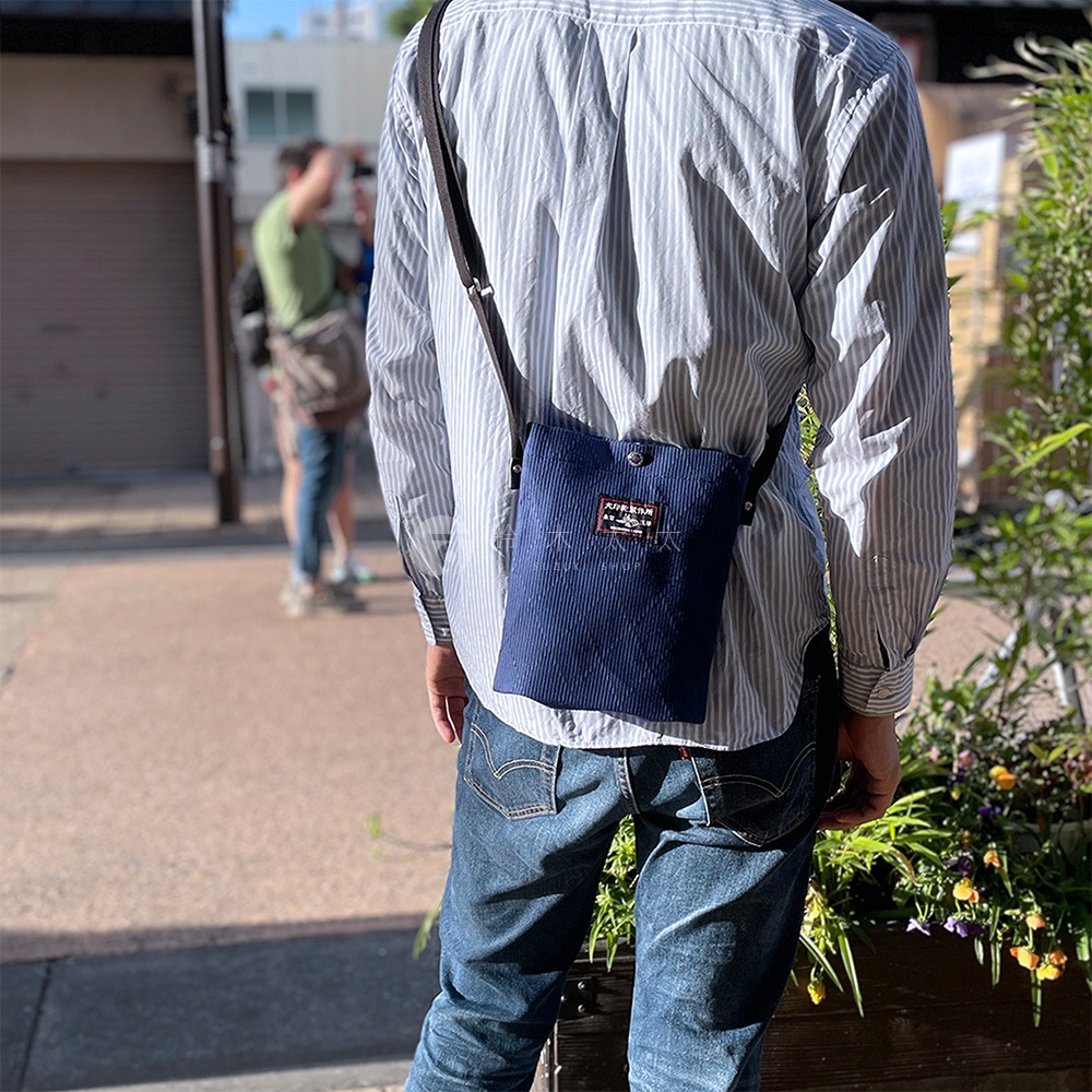 深藍色燈芯絨材質的直立式平板收納側背包，配有黑色斜背帶，包身正面中央有一個方形的品牌布標，由一名男子斜背於身側，男子身穿藍白條紋襯衫與牛仔褲。