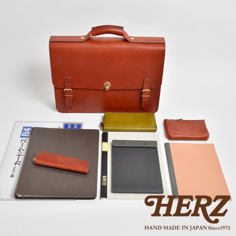 棕色皮革公事包，配有金色鎖扣和搭扣。包旁擺放著綠色長夾、棕色零錢包、灰色文件夾、筆和粉色筆記本。