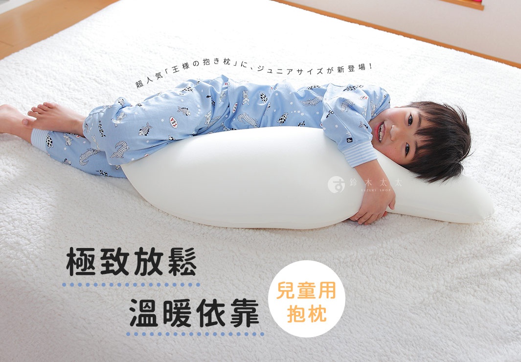 一個男孩抱著一個白色、豆莢形狀的兒童抱枕，抱枕材質看起來柔軟，男孩穿著藍色印有魚圖案的睡衣，躺在鋪有白色絨毛毯子的地板上。