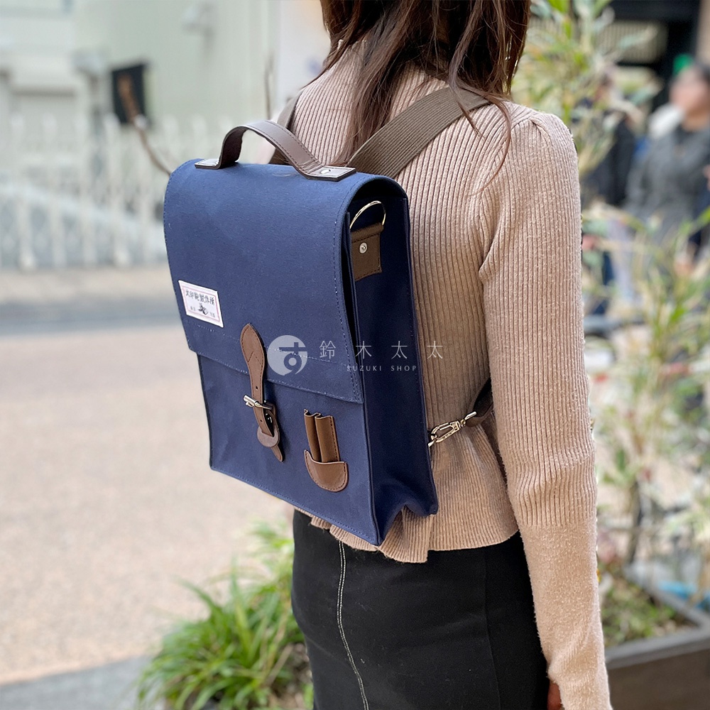 藍色純棉帆布製成的直式側背包，搭配棕色皮革提把、翻蓋束帶與扣環，以及可調節的帆布背帶，背包正面有品牌標籤。