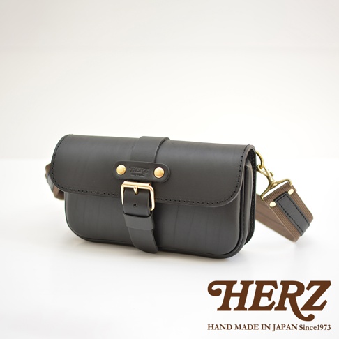 黑色皮革材質的單扣斜背包，正面有品牌標誌和金屬扣環，附有可調節的織帶和皮革肩帶，整體設計簡約經典。