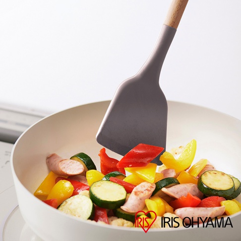 灰色矽膠鍋鏟正在翻炒鍋中的香腸、櫛瓜、紅黃甜椒等食材，鍋鏟手柄為木質。