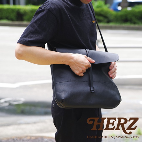 一手拿著黑色皮革包的男子，包包上有細扣帶設計，下方有HERZ品牌標誌。