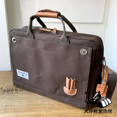 深棕色純綿帆布商務公事包，B4尺寸，配有皮革提把與肩帶，正面有鈕扣裝飾及品牌標籤，簡約實用。