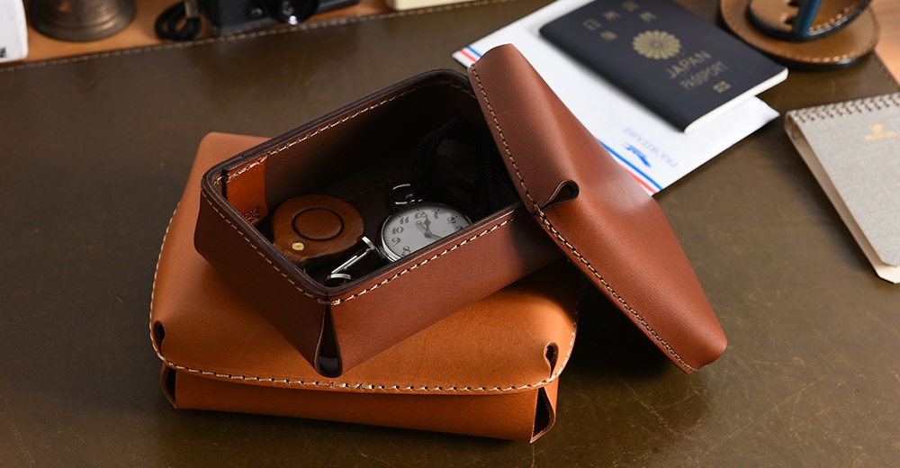 皮革小物收納盒，展示於桌面，盒內有懷錶、鑰匙圈等小物，盒身與盒蓋皆為棕色系皮革，縫線清晰。