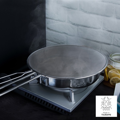 一個銀色的金屬網狀防噴濾網，帶有三個金屬手柄，覆蓋在一個銀色金屬鍋的上方，鍋架在一個銀色的爐子上，背景是黑白相間的牆壁，右側有一個裝有檸檬片的玻璃罐。