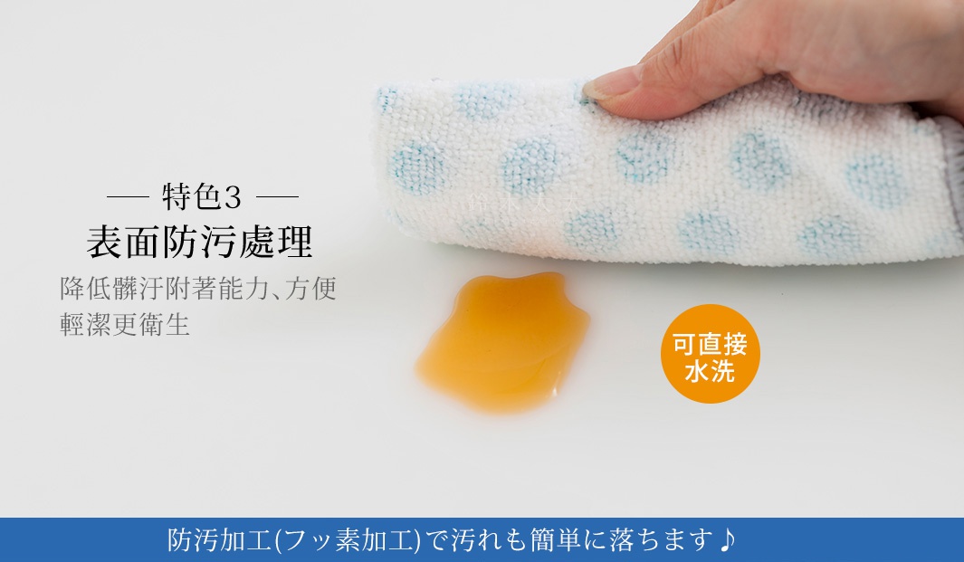 手指拿著一塊白色帶有藍色圓點的毛巾，擦拭桌面上的一灘黃色液體，旁邊有寫著「可直接水洗」的橘色圓形標籤，並有文字說明「表面防污處理」，「降低髒污附著能力、方便輕潔更衛生」。