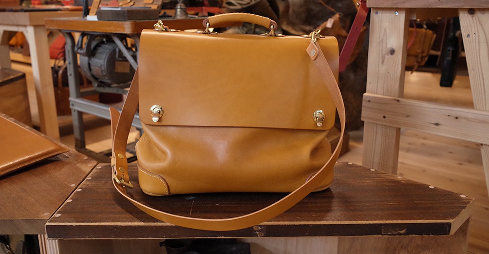 職人手作皮革旅行商務兩用波士頓包，呈現自然原色皮革，款式簡潔，配有頂部提把及可拆卸斜背帶，金色金屬扣環裝飾，質感厚實，適合商務與旅行等多種場合。