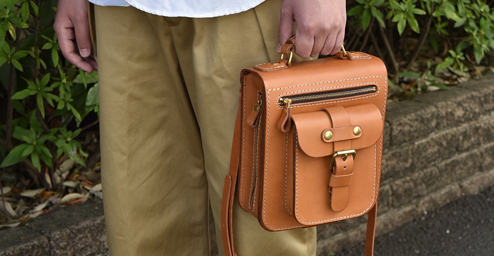 棕色皮革製成的手提斜背包，具備立體前袋、拉鍊開合及金屬扣環，整體呈現職人手作質感。