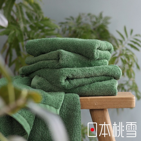 日本桃雪精梳棉綠色浴巾堆疊在木凳上，背景為綠葉植物，右下角有日文品牌標誌。
