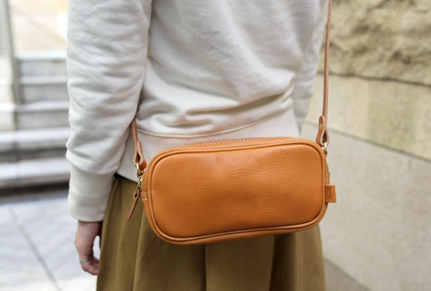 一個棕褐色皮革製成的長方形隨身小包，配有細長皮革肩帶和金色拉鍊。包包表面有明顯的皮革紋理和手工縫線。