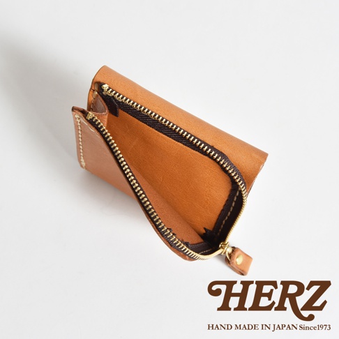 一個由日本手工皮革品牌HERZ設計的淺棕色皮革短夾，採用簡約的方形拉鍊設計，呈現皮革自然紋理與精緻的車縫細節，右下角印有品牌名稱與手工製造年份字樣。