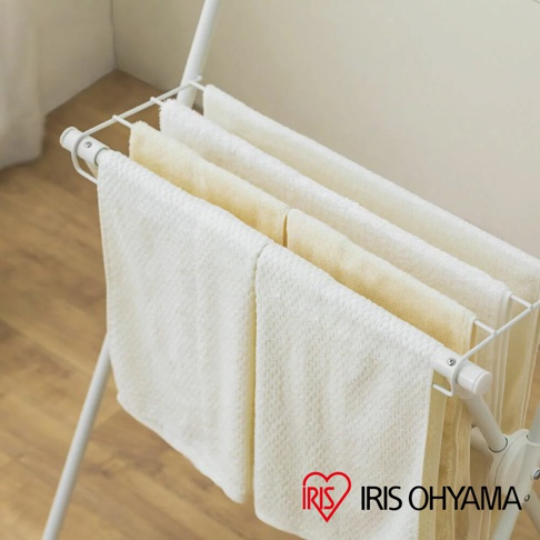 白色金屬X型曬衣架，架上掛有數條米白色及淺黃色毛巾，架子下方有IRIS OHYAMA品牌標誌。