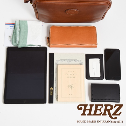 職人手作皮革可拆式扣環萬用手提包 S-11，以棕色皮革製成，搭配可拆卸的扣環。包內物品包含一個棕色長夾、一個黑色平板電腦、一本淺色書籍、一張卡夾、一個黑色手機及一條綠色毛巾。