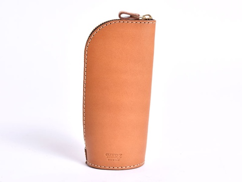 一個棕褐色皮革製成的直立式筆袋，頂部有拉鍊和皮革拉環，側面縫有白色縫線，底部可見品牌標誌「HERZ」及「MADE IN JAPAN」字樣。