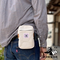 米白色石洗帆布斜背手機包，附有棕色皮革掛環，包身中央有粉色方形標籤，印有黑白圖案和日文字樣。