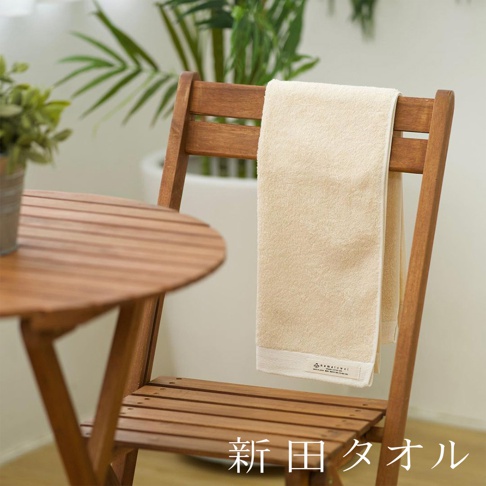 米色有機棉柔無漿生浴巾，懸掛在木椅上，旁邊是木桌和室內綠植。
